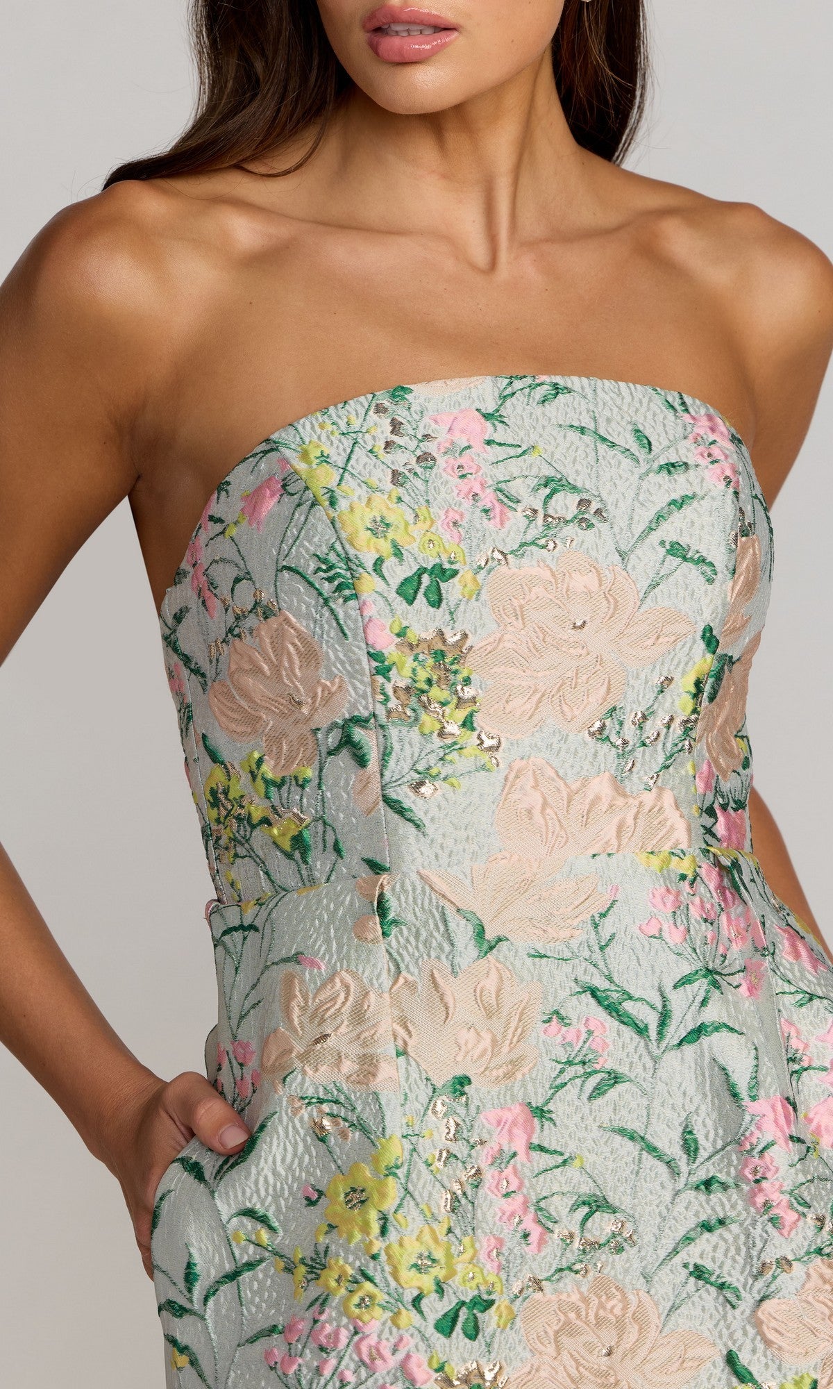 A woman models the Mac Duggal 13223, a tea-length party dress adorned with a pastel floral brocade design and delicate green, pink, and yellow embroidery. The strapless silhouette features practical pockets, all set against a plain neutral background that highlights the elegance of this creation.